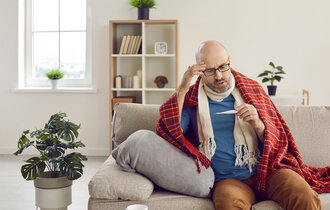 Ein Mann, der auf einem Sofa in einem Wohnzimmer sitzt. Er Hat eine Decke und einen Schal um sich und schaut auf ein Fieberthermometer.