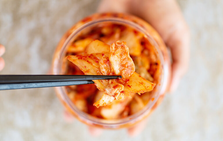 Kimchi in einem Glas von oben. Zwei chinesische Stäbchen holen ein Stück heraus. 