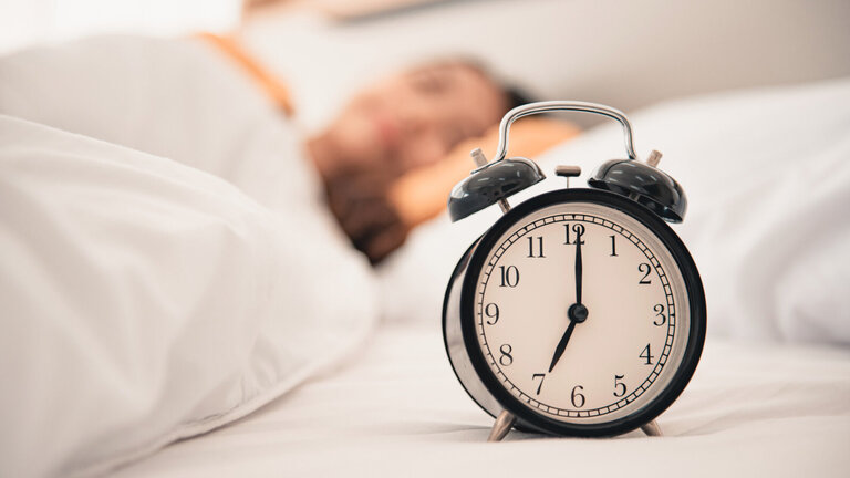 Ein großer Wecker, der 7 Uhr zeigt. Im Hintergrund unscharf eine schlafende Person.